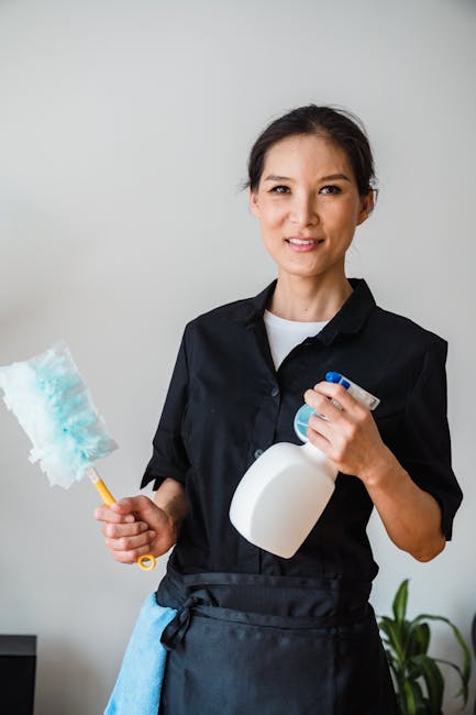 A professional cleaner wearing a white protective suit, orange gloves, and white sneakers is performing surface cleaning on a large gray tiled floor in a modern living room. The cleaner is using a vac