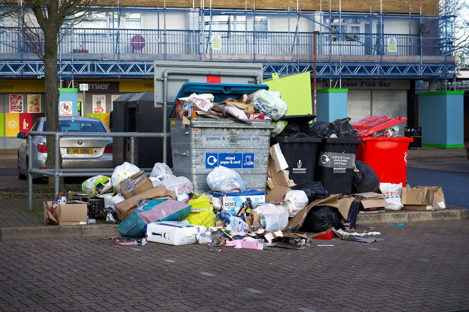 A roadside waste disposal area featuring a large metal mixed paper and cardboard recycling bin overflowing with crumpled paper, cardboard boxes, and plastic bags. Surrounding the bin are additional bl