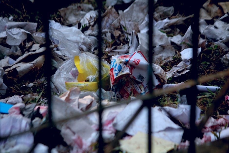 A roadside waste disposal area featuring a large metal mixed paper and cardboard recycling bin overflowing with crumpled paper, cardboard boxes, and plastic bags. Surrounding the bin are additional bl